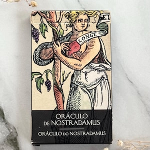 May include: A tarot card deck featuring a vintage illustration of a woman holding a tree branch with grapes. The card has the word "LONGE" on a banner. The title "ORÁCULO DE NOSTRADAMUS" is printed on a black band at the bottom.