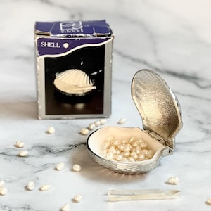 May include: A silver-colored, shell-shaped container filled with small, off-white pearls. A small box with the word "SHELL" is visible. Scattered pearls and a small tool are also present.
