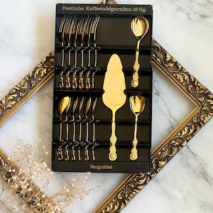 May include: A 15-piece gold-colored flatware set in a black box. The set includes forks, spoons, and a cake server. The box is labeled "Festliche Kaffeetafelgarnitur, 15-tlg." and "Vergoldet."