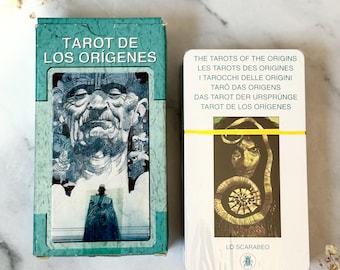Puede incluir: Un juego de cartas de tarot turquesa y blanco. La caja dice "TAROT DE LOS ORÍGENES" con una ilustración detallada. La baraja de cartas dice "THE TAROTS OF THE ORIGINS" en varios idiomas. La baraja presenta una banda amarilla.