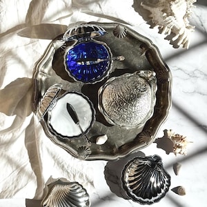 Könnte beinhalten: Eine Sammlung von silberfarbenen, muschelförmigen Servierschalen und einem Tablett. Eine Schale hat einen leuchtend blauen Innenraum, eine andere enthält eine weiße Schale mit einem kleinen Messer. Die Anordnung umfasst eine große Muschel und ist auf einem weißen Tuch platziert.