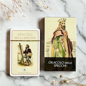 Può includere: Un mazzo di carte dei tarocchi "Oracolo Degli Specchi". La scatola delle carte presenta un'illustrazione di un re con una spada. Il mazzo di carte mostra un artista che dipinge su un cavalletto. Il testo "Oracolo Degli Specchi" è stampato.