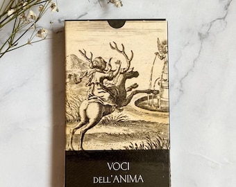 May include: A rectangular box with a vintage illustration of a figure riding a deer, with a fountain in the background. The box has the words "Voci Dell'Anima" printed on the bottom in black. The box is set against a white and gray marble surface.