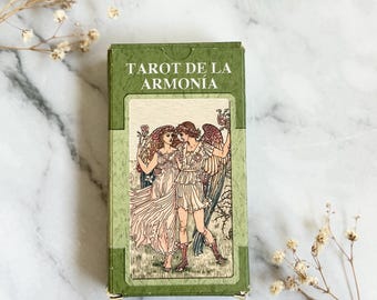 Op de afbeelding: Een groene tarotkaartdoos met de woorden "TAROT DE LA ARMONIA" gedrukt op de bovenkant. De doos heeft een illustratie van drie figuren in vloeiende gewaden. De achtergrond is een marmeren oppervlak met gedroogde bloemen.