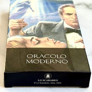May include: A tarot card box featuring a vintage illustration of a man in a suit holding a pendant over a woman. The box has the words "ORACLO MODERNO" and the Lo Scarabeo logo. The box is black with a white marble background.