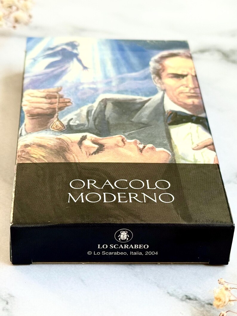 May include: A tarot card box featuring a vintage illustration of a man in a suit holding a pendant over a woman. The box has the words "ORACLO MODERNO" and the Lo Scarabeo logo. The box is black with a white marble background.