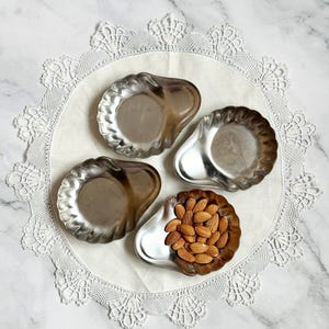 May include: Four small, silver-colored, pear-shaped serving dishes with scalloped edges are arranged on a white, lace-edged doily. One dish is filled with almonds. The doily rests on a marble surface.