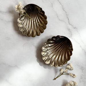 May include: Two metallic gold seashell-shaped dishes with a ribbed design. The dishes are placed on a white marble surface and decorated with small white flowers.