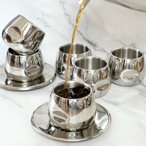 Pode incluir: Um conjunto de xícaras e pires de café expresso em aço inoxidável. O café está sendo despejado em uma xícara de uma jarra de aço inoxidável combinando. As xícaras têm um design curvo exclusivo e estão em uma superfície de mármore branco.