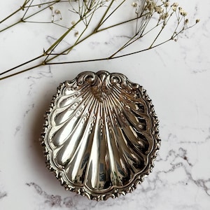 May include: A silver-colored, shell-shaped dish with intricate detailing. The dish has a scalloped edge and a reflective surface. Delicate white flowers and stems are in the background, creating a contrast with the metallic dish.