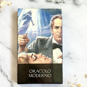 May include: A tarot card deck box featuring a vintage illustration of a man holding a pendant over a woman, with a woman in a flowing gown in the background. The box has the words "ORACLO MODERNO" in white on a black background.