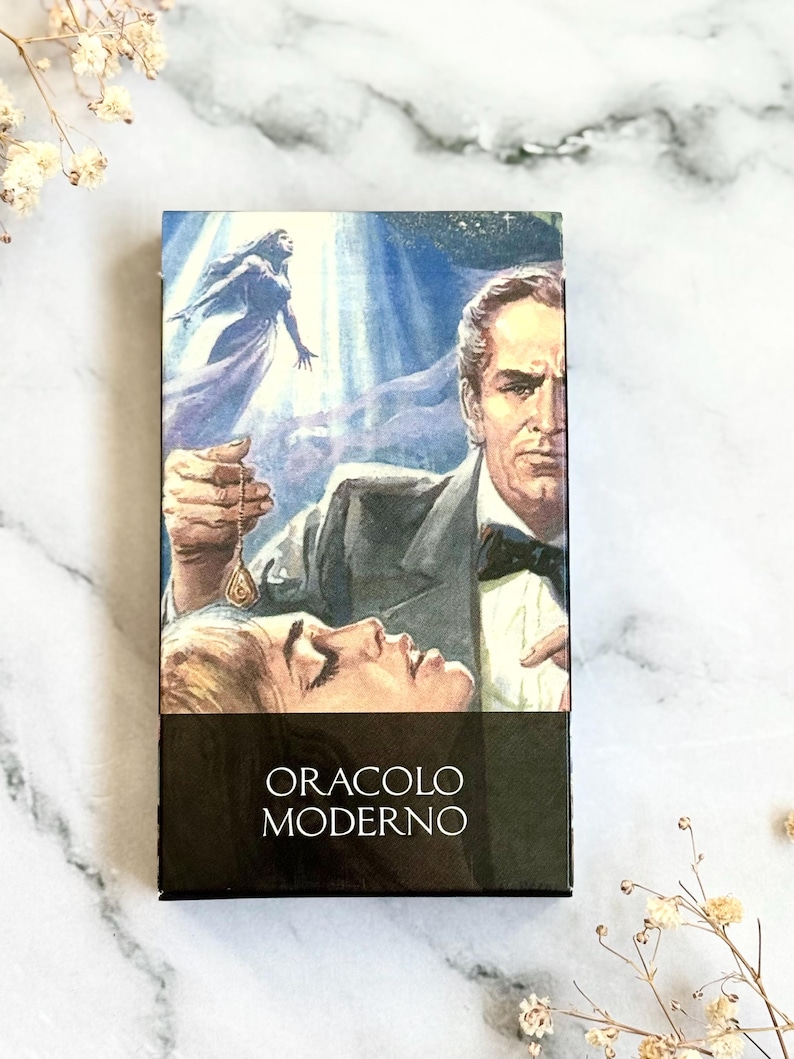 May include: A tarot card deck box featuring a vintage illustration of a man holding a pendant over a woman, with a woman in a flowing gown in the background. The box has the words "ORACLO MODERNO" in white on a black background.