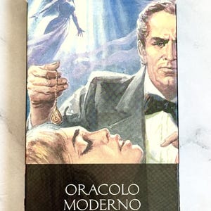 May include: A tarot card deck box featuring a vintage illustration. The artwork depicts a man holding a pendulum over a woman, with a floating figure in the background. The text "ORACLO MODERNO" is printed on the bottom.