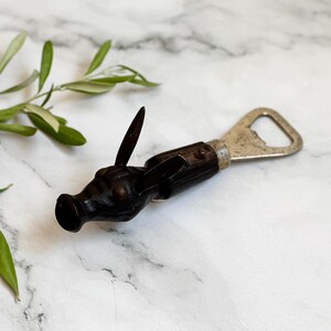 Puede incluir: Un abridor de botellas marrón oscuro con un diseño decorativo de cabeza de animal. El abridor tiene un abridor de botellas de metal y una pequeña cuchilla. El abridor está sobre un fondo de mármol blanco. El diseño es de caballo.