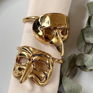 Può includere: Due anelli per tovaglioli in metallo dorato a forma di maschera. Le maschere hanno un naso lungo e appuntito e buchi per gli occhi.