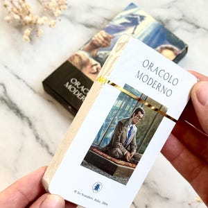 May include: A deck of oracle cards titled "ORACLO MODERNO" in white and gold. The card features an image of a man in a suit. The deck is held in a person's hands. The box in the background has the same title.
