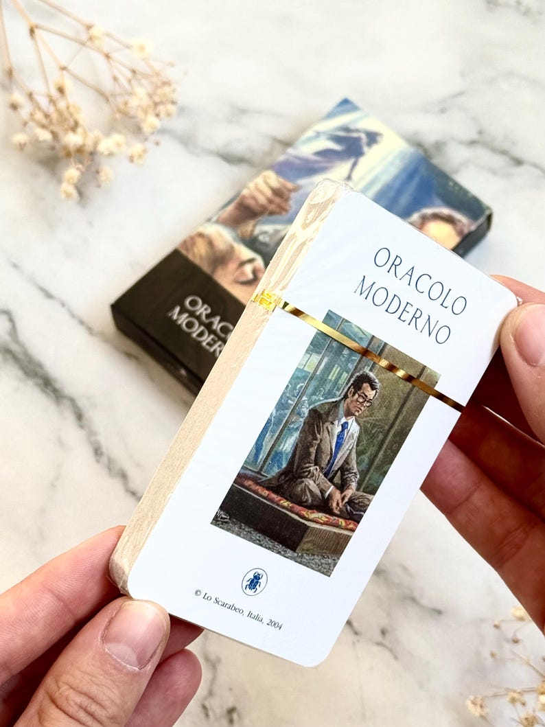 May include: A deck of oracle cards titled "ORACLO MODERNO" in white and gold. The card features an image of a man in a suit. The deck is held in a person's hands. The box in the background has the same title.