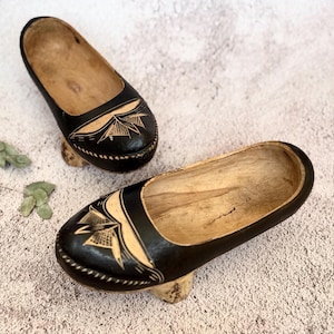 May include: A pair of wooden clogs with black painted tops and intricate white designs. The clogs have a rounded toe and a low heel.