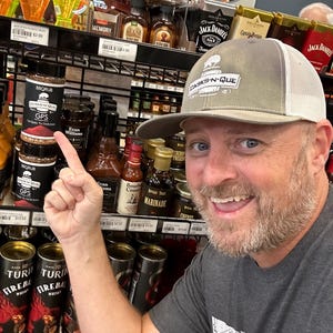 May include: A man wearing a grey and white baseball cap with the text "CASKS-N-QUE" pointing to a small jar of BBQ rub with the text "GPS" and "CASKS-N-QUE" on the label. The jar is on a shelf with other jars and bottles of various sauces and marinades.