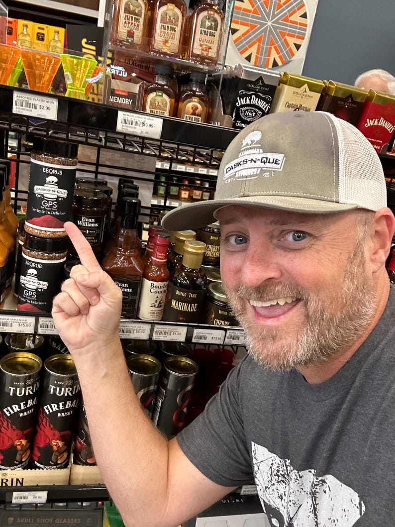 May include: A man wearing a grey and white baseball cap with the text "CASKS-N-QUE" pointing to a small jar of BBQ rub with the text "GPS" and "CASKS-N-QUE" on the label. The jar is on a shelf with other jars and bottles of various sauces and marinades.