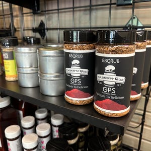 May include: A shelf display of various spice jars and tins. The jars are labelled "BBQRUB" and "CASKS-N-QUE" with the text "GPS Salt Pepper Garlic Navigate To Delicious".