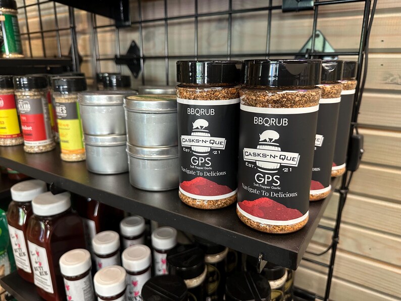 May include: A shelf display of various spice jars and tins. The jars are labelled "BBQRUB" and "CASKS-N-QUE" with the text "GPS Salt Pepper Garlic Navigate To Delicious".