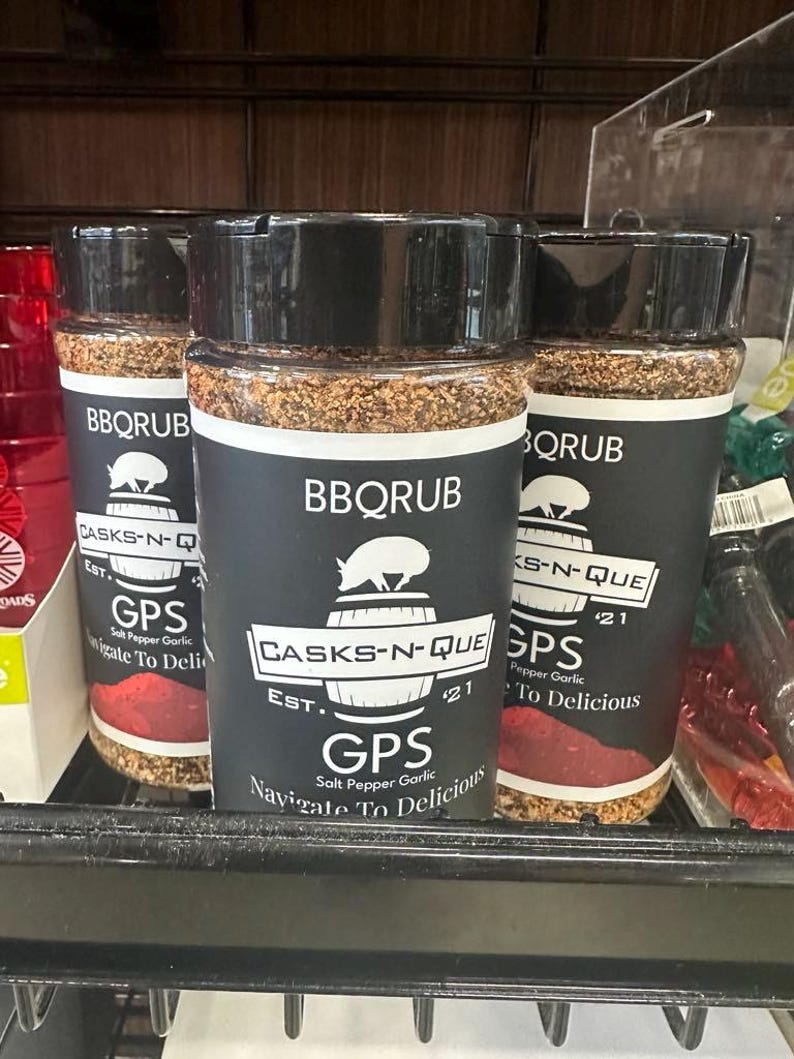 May include: Three glass jars of BBQ rub with black lids and labels. The labels have a pig and a barrel logo with the text "Casks-N-Que Est. '21 GPS Salt Pepper Garlic Navigate To Delicious" on the front.