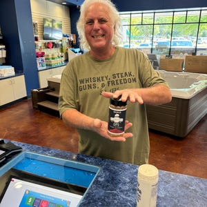 May include: A man wearing a green t-shirt with the text "Whiskey, Steak, Guns, & Freedom" is holding a small bottle of seasoning labelled "BORUB GPS Garlic Pepper Seasoning".