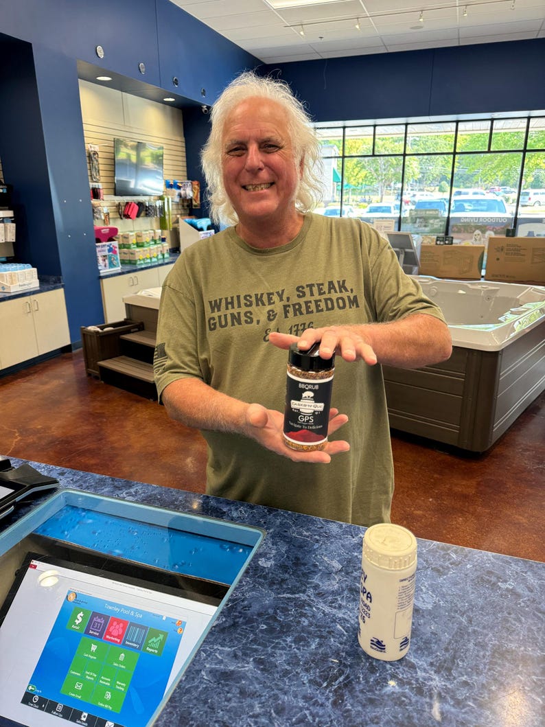 May include: A man wearing a green t-shirt with the text "Whiskey, Steak, Guns, & Freedom" is holding a small bottle of seasoning labelled "BORUB GPS Garlic Pepper Seasoning".