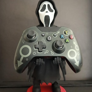 Könnte beinhalten: Eine Figur von Ghostface aus den Scream-Filmen, die einen dunkelgrünen Xbox-Controller hält. Der Controller hat farbige Knöpfe und ein Design mit Symbolen. Die Figur steht auf einem roten Sockel.