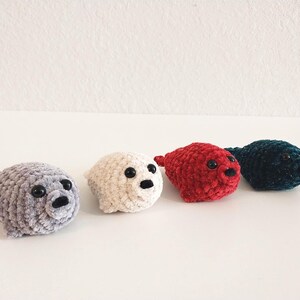 May include: Four small, plush crochet seal toys arranged in a row. The seals are colored light gray, cream, red, and dark teal. Each seal features a rounded body, small flippers, a snout with a black nose, and two shiny black plastic eyes.