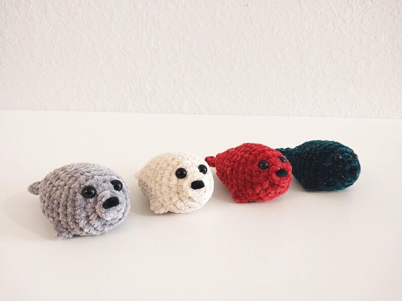 May include: Four small, plush crochet seal toys arranged in a row. The seals are colored light gray, cream, red, and dark teal. Each seal features a rounded body, small flippers, a snout with a black nose, and two shiny black plastic eyes.