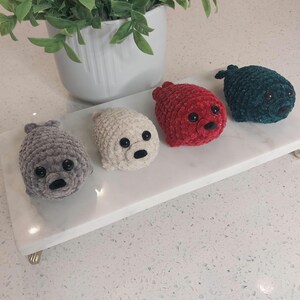 May include: Four small, plush, crocheted seal figures arranged in a row. The seals are colored grey, cream, red, and dark teal. Each seal features a rounded body, two small flippers, a black nose, and two round, black plastic safety eyes.