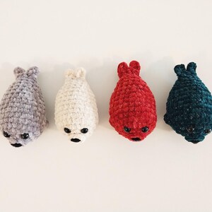 May include: Four small, handmade crochet seal plushies arranged in a row. The seals are colored light gray, cream, bright red, and dark teal. Each plush features a textured yarn finish, two small black plastic eyes, and a black stitched nose.