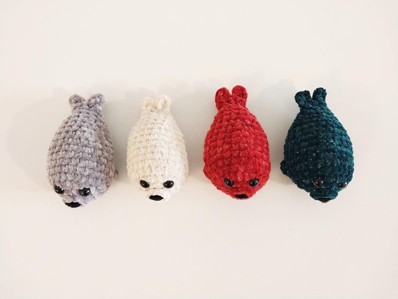 May include: Four small, handmade crochet seal plushies arranged in a row. The seals are colored light gray, cream, bright red, and dark teal. Each plush features a textured yarn finish, two small black plastic eyes, and a black stitched nose.