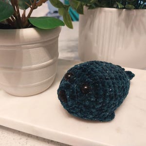 May include: A small, dark teal plush seal toy crafted from soft, textured yarn. The seal features two round, dark brown plastic eyes and a small, dark brown nose. The material has a subtle, shimmering quality throughout the crocheted surface.