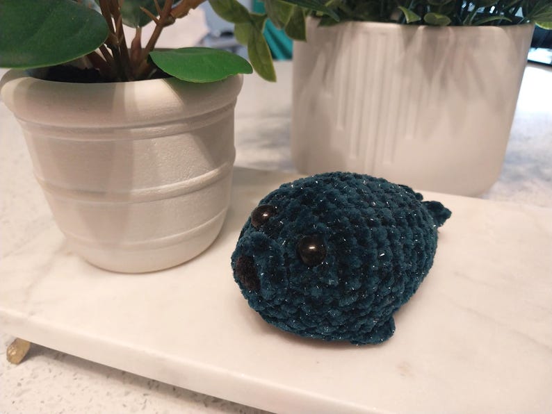 May include: A small, dark teal plush seal toy crafted from soft, textured yarn. The seal features two round, dark brown plastic eyes and a small, dark brown nose. The material has a subtle, shimmering quality throughout the crocheted surface.
