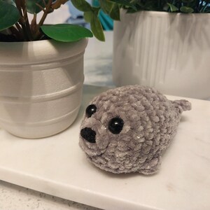May include: A small, handmade crochet seal plush toy crafted from soft, light gray velvet yarn. The seal features two round, glossy black plastic safety eyes and a small, triangular black embroidered nose, with a rounded body and flippers.