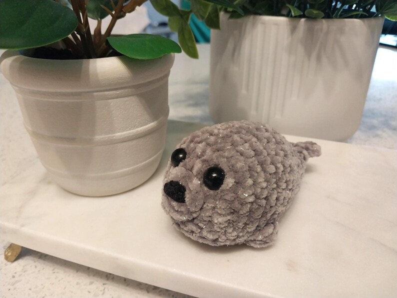 May include: A small, handmade crochet seal plush toy crafted from soft, light gray velvet yarn. The seal features two round, glossy black plastic safety eyes and a small, triangular black embroidered nose, with a rounded body and flippers.