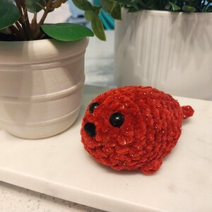 May include: A small, round, crocheted plush toy in a vibrant red color with a soft, fuzzy texture. It features two large, glossy black plastic eyes and a small, triangular black nose, resembling a seal with a compact, stuffed body shape.