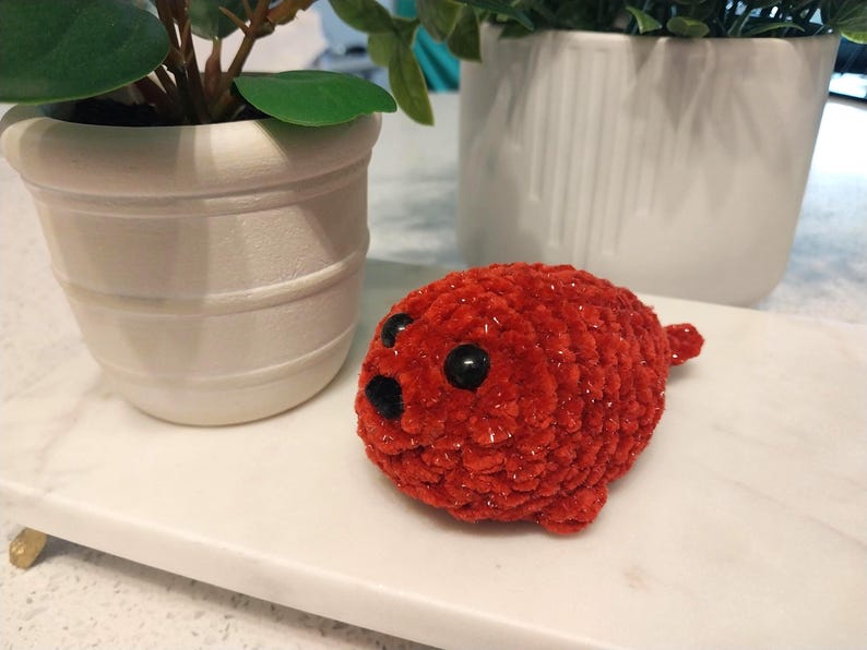May include: A small, round, crocheted plush toy in a vibrant red color with a soft, fuzzy texture. It features two large, glossy black plastic eyes and a small, triangular black nose, resembling a seal with a compact, stuffed body shape.