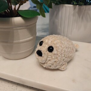May include: A small, cream-colored crocheted seal figurine with a textured, fuzzy yarn surface. The seal features two round, glossy black plastic eyes and a small, triangular black stitched nose, giving it a rounded, compact appearance.
