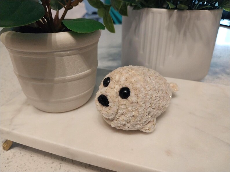 May include: A small, cream-colored crocheted seal figurine with a textured, fuzzy yarn surface. The seal features two round, glossy black plastic eyes and a small, triangular black stitched nose, giving it a rounded, compact appearance.