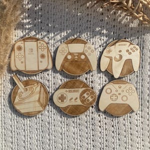 May include: Six wooden coasters featuring different video game controllers. The coasters are circular and have a brown wood grain finish. The controllers include a Nintendo Switch, a Playstation 5, a Super Nintendo, an Xbox, a Sega Genesis, and a Nintendo 64.