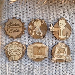 May include: Six wooden Fallout-themed coasters. The coasters feature various designs, including the Nuka Cola logo, a robot, a character giving a thumbs up, and a computer. The coasters are made of light-coloured wood.