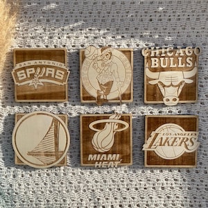 Peut inclure: Six sous-verres en bois avec des logos d'équipes de la NBA. Les sous-verres sont carrés et ont une finition en bois brun clair. Les logos sont gravés en blanc. Les logos sont pour les San Antonio Spurs, les Boston Celtics, les Chicago Bulls, les Golden State Warriors, le Miami Heat et les Los Angeles Lakers.