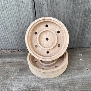 May include: Two unfinished wooden coasters with a circular design. Each coaster has a raised outer rim and a central recessed area with several small holes. The coasters are made of light-colored wood and are stacked on a weathered wooden surface.