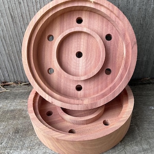May include: Two-piece, reddish-brown wooden item. The top piece is a shallow, circular lid with a central raised ring and several small holes. The bottom piece is a matching base. The wood grain is visible, and the items appear to be handmade.