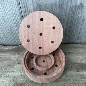 May include: A wooden, circular box with a lid. The box and lid are made of a light brown wood and feature several small holes. The lid is slightly ajar, revealing the interior of the box, which has concentric circular tiers.