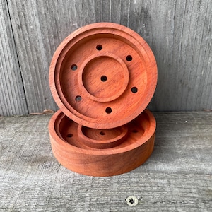 May include: Two reddish-brown wooden coasters. Each coaster has a circular design with a raised outer rim and a central, smaller raised circle. Both coasters have several small holes. The coasters are stacked on top of each other.